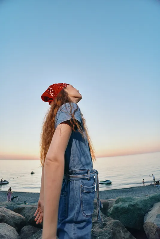Lifestyle woman in modern boho-western denim street style with a red bandana, enjoying serene ocean view in warm film color tones at sunset.