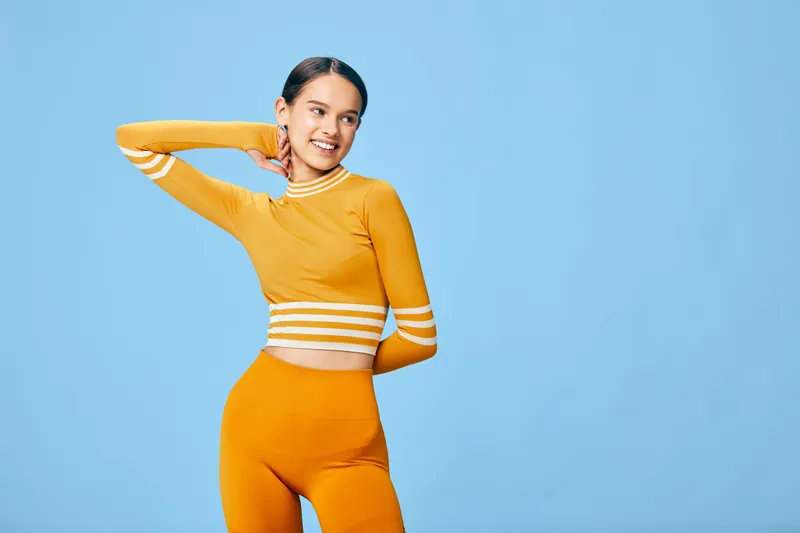 Woman in bright orange activewear poses confidently in a studio, showcasing fitness style, flexibility, and energy against a clean blue background with a cheerful vibe.