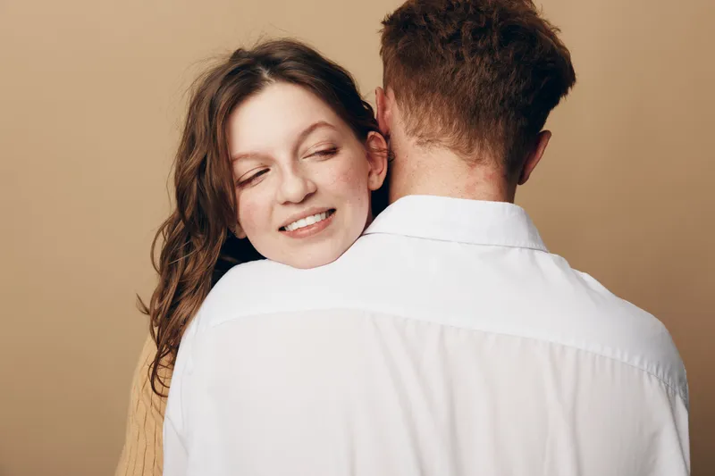Smiling woman with closed eyes hugs man wearing white shirt from behind. Intimate moment of affection and love between couple on beige background. Concept of relationship and tenderness.