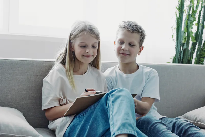 Two children sitting on a couch, focused on drawing with a pencil on a clipboard. Natural light and casual clothing create a relaxed home atmosphere with creative play.