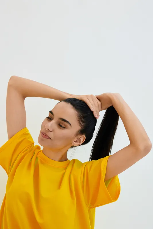 young woman tying her long dark hair wearing a bright yellow shirt with a calm expression, isolated on white background, casual and natural pose
