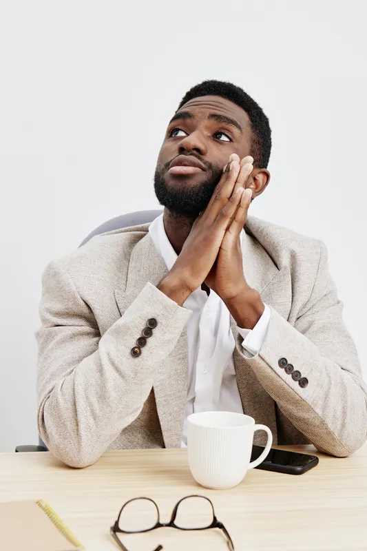 Thoughtful young man with dark skin, short curly hair, wearing beige blazer and white shirt, sitting at desk with coffee mug, smartphone, and glasses, looking upward with hands clasped, studio shot. P