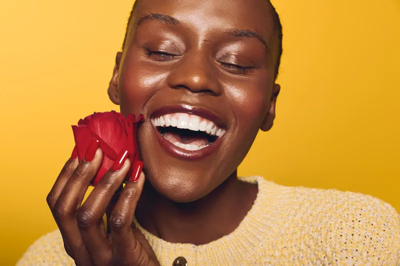 Close-up of smiling African woman with dark skin holding red rose near face, eyes closed, wearing yellow sweater on yellow background. Concept of happiness, beauty, and natural skincare.