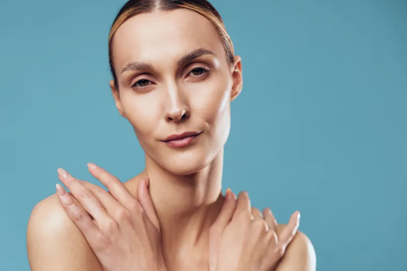 Young woman with clear skin stands against a blue background, touching her shoulders in a calm pose that suggests skincare and self-care. Soft, even lighting adds warmth and a polished beauty