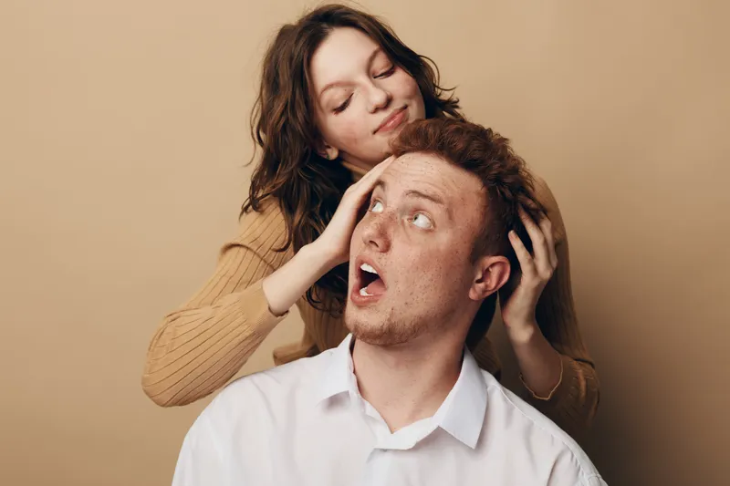 Young woman playfully covers surprised man eyes with hands in beige studio. Concept of fun, relationship, trust, and playful interaction between couple with casual clothing and natural expressions.