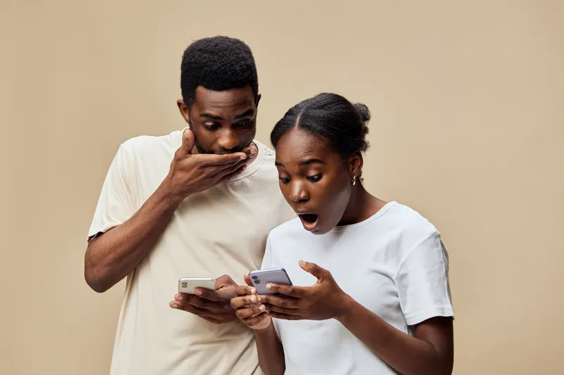 Excited young couple reacting to shocking news on smartphones, showcasing surprise and curiosity in neutral setting