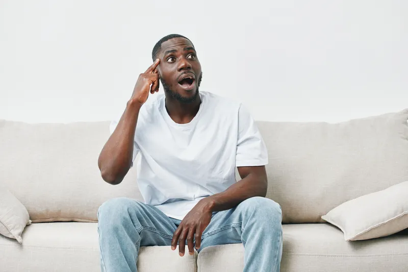 Surprised young black man in casual outfit sitting on sofa, showcasing emotions and thoughts, expressing surprise and contemplation in a modern living room setting