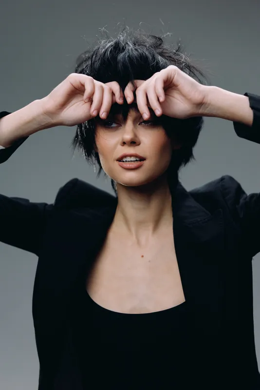 confident woman with short black hair posing with hands near face in black outfit on gray background