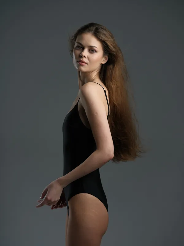 Young woman with long hair wearing black bodysuit standing against gray background in studio lighting.