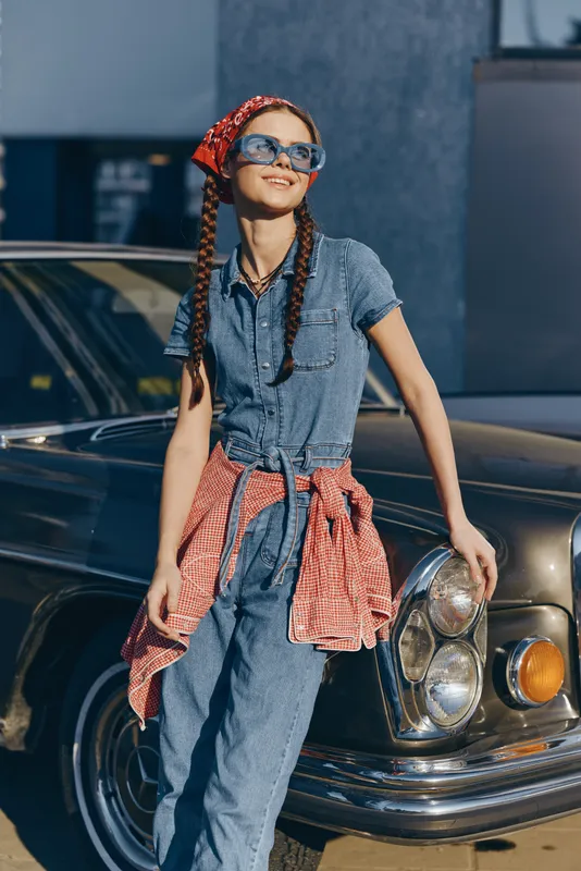 Lifestyle woman in modern boho-western denim street style wearing vintage sunglasses and a red scarf, standing confidently by a classic car in bright film color tones.