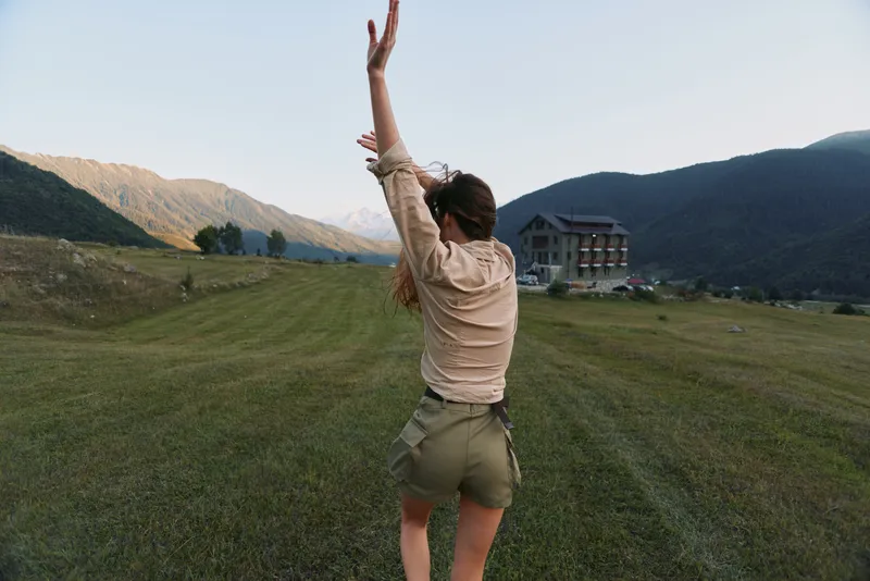 Woman in an outdoor field raises arms in celebration, back view, wearing beige shirt and shorts, open landscape with mountains, capturing a joyful lifestyle moment in nature