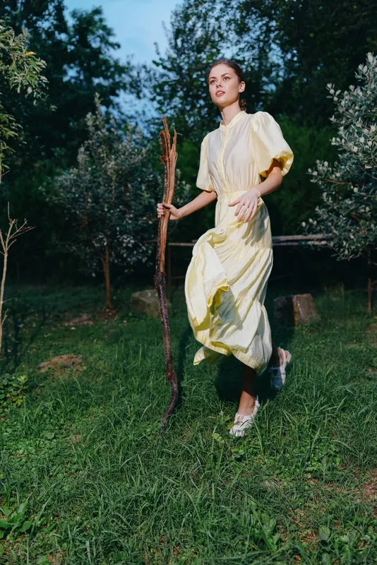 Woman in a light yellow dress runs through a grassy garden, holding a wooden stick, enjoying a sunny outdoor moment amid trees and fresh air.