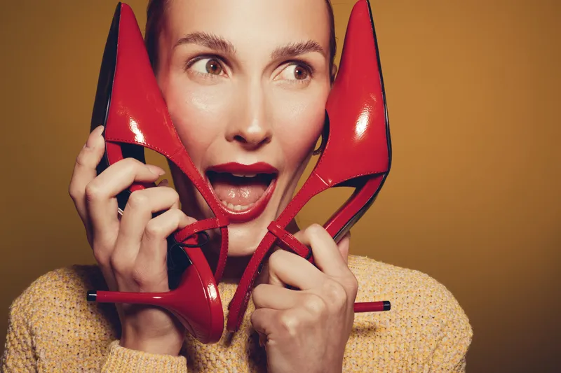 Excited woman holds red high heels near face with open mouth expression. Studio portrait on brown background. Concept of fashion, shoes, style, and female emotions.
