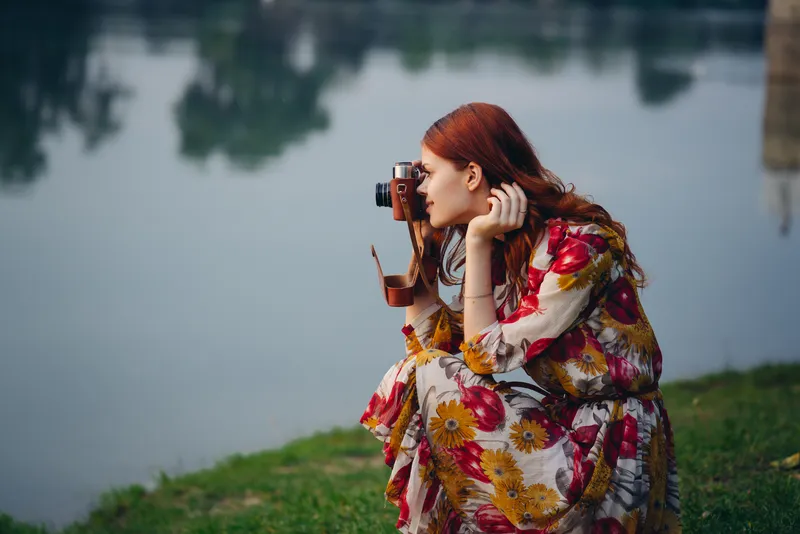 Женщина в платье с фотоаппаратом на природе, снимающая реку в качестве хобби