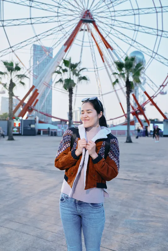 Smiling woman in a brown jacket and jeans stands in a sunlit outdoor plaza with a towering ferris wheel behind, capturing casual urban style, travel vibe, and street fashion in a candid moment