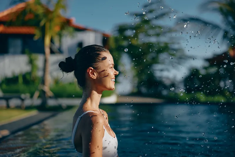Smiling woman in white swimsuit enjoys water splashes near poolside with tropical palm trees and house in background on sunny day, lifestyle concept outdoors.