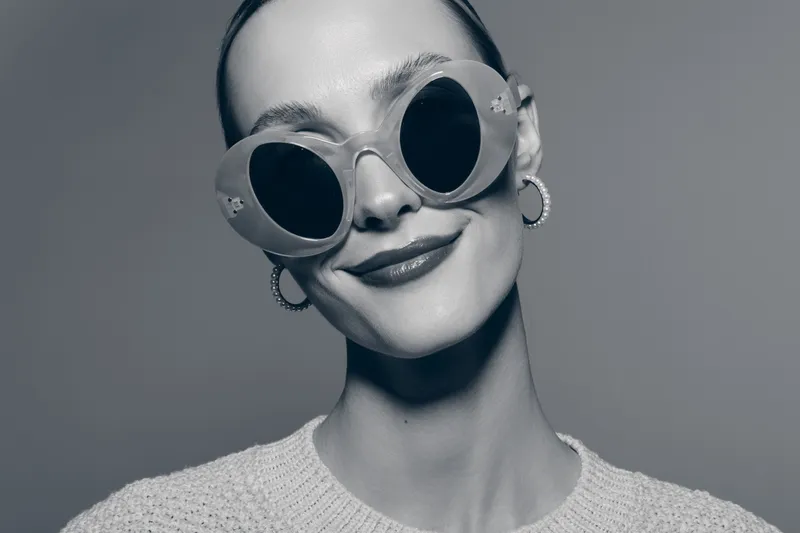 Smiling white woman with pale skin wearing round sunglasses and hoop earrings poses in knitted sweater on grey background. Concept for fashion and accessories.