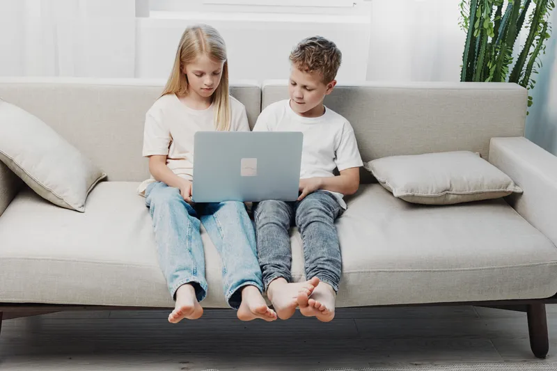 Two barefoot children sitting on a modern sofa using a laptop together, focused on the screen in a bright living room with soft cushions and natural light.
