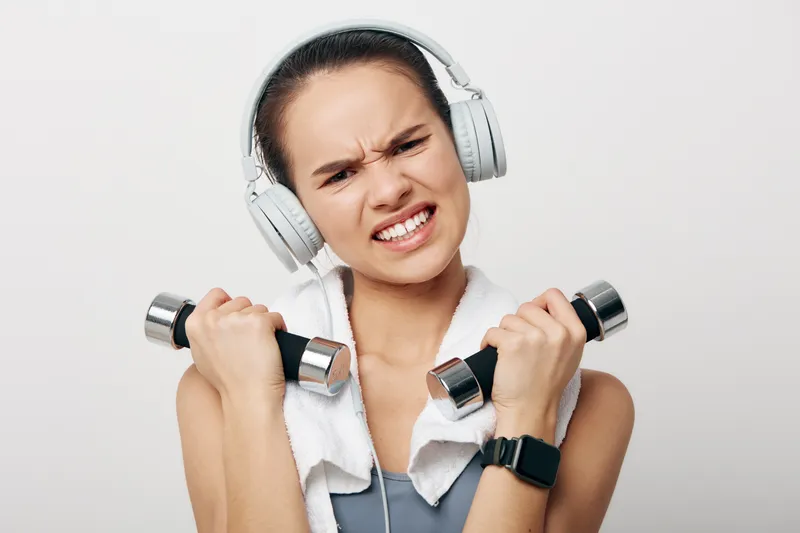 Woman fitness enthusiast lifts dumbbells with a towel around her neck and headphones, showing intense focus during a dynamic workout in a studio setting.