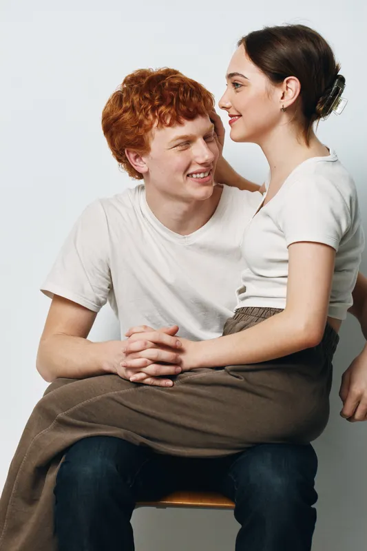 Young couple sitting together smiling and looking at each other in casual clothing with relaxed mood and friendly interaction on white background.