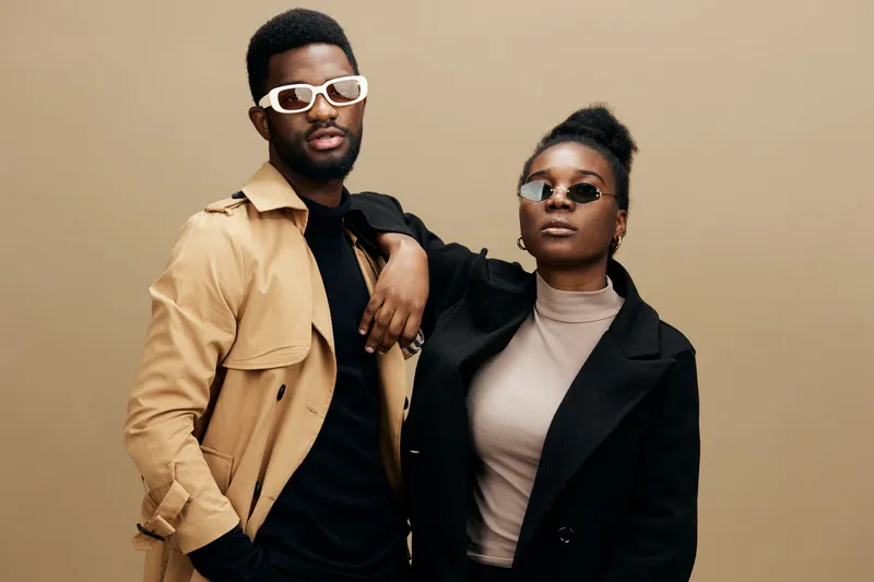 Stylish young couple in trendy outfits poses confidently against a neutral background, showcasing modern fashion and elegance with sunglasses and smiles