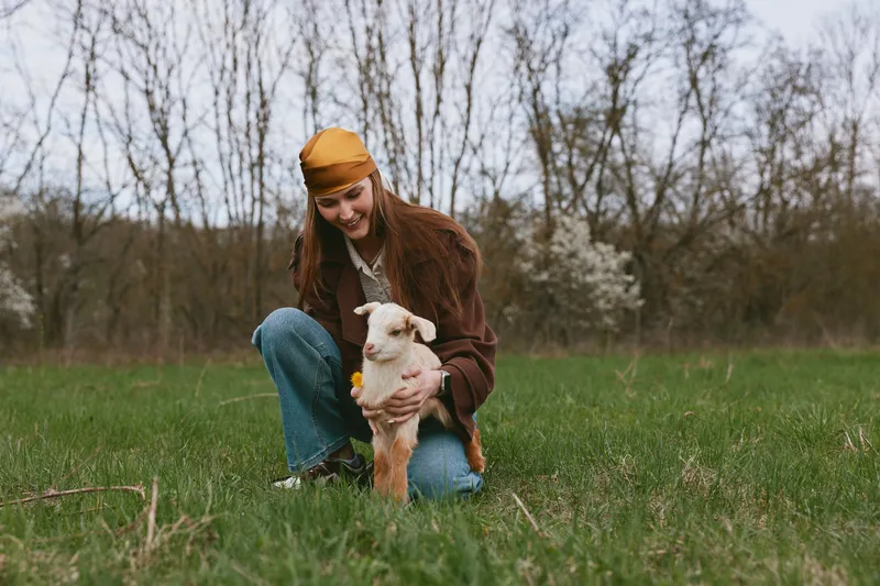 Young woman outdoors with a goat kid, kneeling on grass, smiling, wearing a mustard hat and brown jacket, gentle interaction, natural lifestyle moment in a park setting, daytime light, wholesome scene
