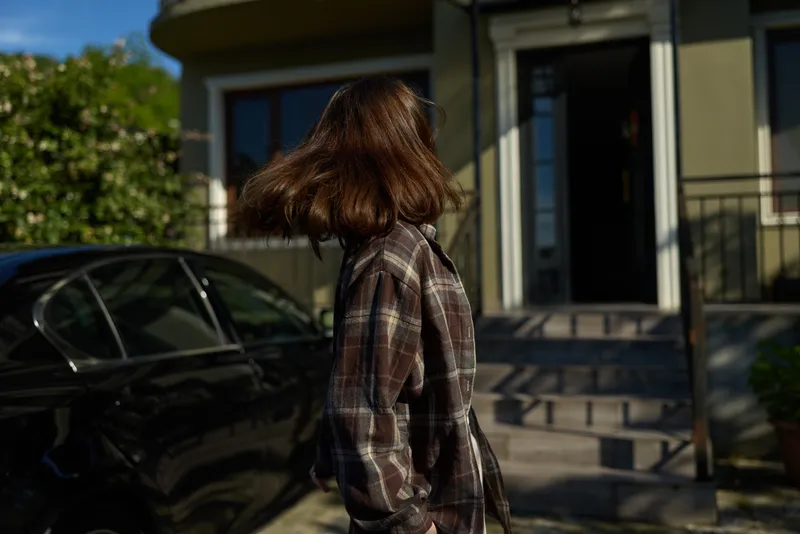A woman with shoulder length hair in a plaid shirt stands outside a modern home, back to the camera. A casual lifestyle moment exploring everyday resilience and quiet confidence