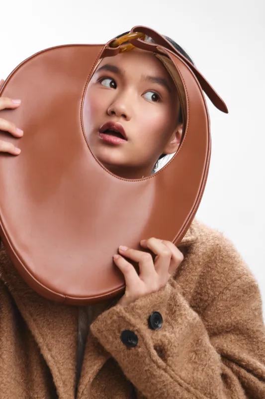 Asian woman holds a brown circular prop near her face, wearing a warm woolen coat and glasses perched on her head. She gazes upward with a curious, surprised expression.