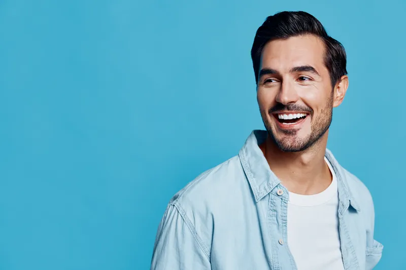 Smiling cheerful young man wearing denim shirt looks at the camera against a solid blue background, radiating positive energy and joy. Lifestyle concept.