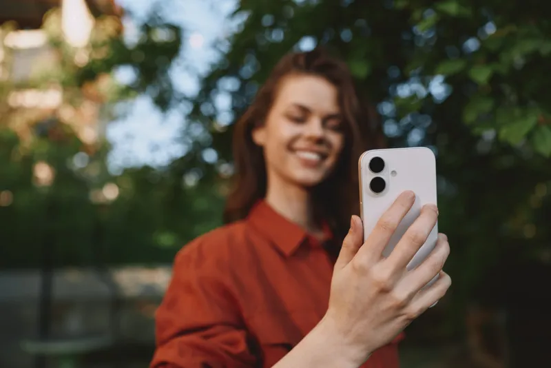 Smiling woman in an orange shirt taking a selfie outdoors, with trees in the background. Casual lifestyle moment showing warmth, natural light, and confident self expression