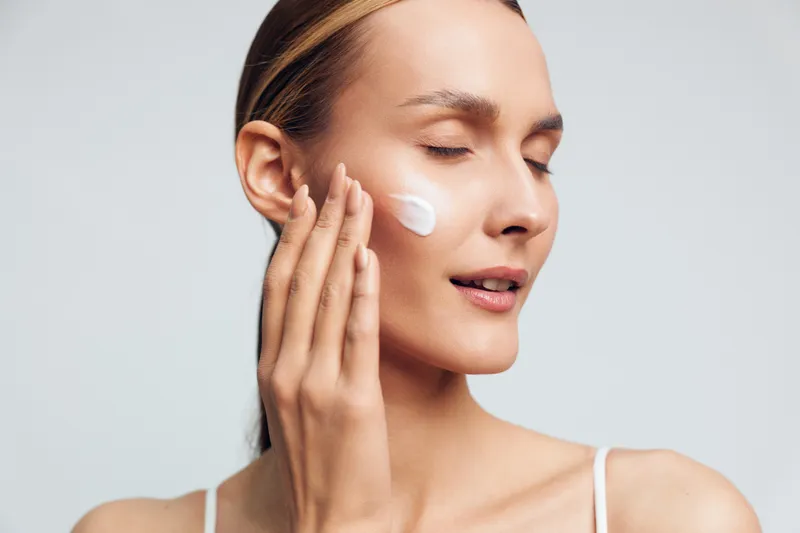 Young woman applying face cream with closed eyes indoors, smooth skin, gentle expression, skincare routine, natural light, minimal background, beauty concept.