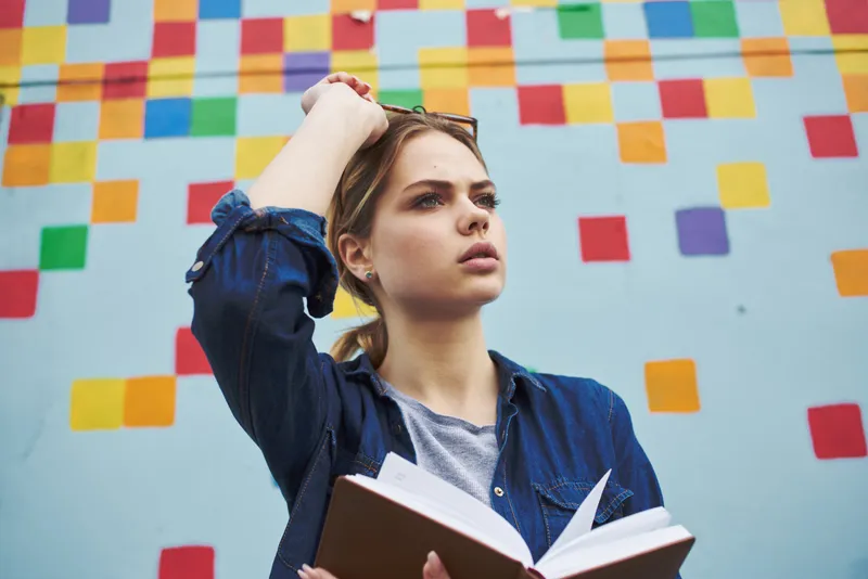 Студентка в рубашке и очках держит в руках книгу на улице возле граффити на стене