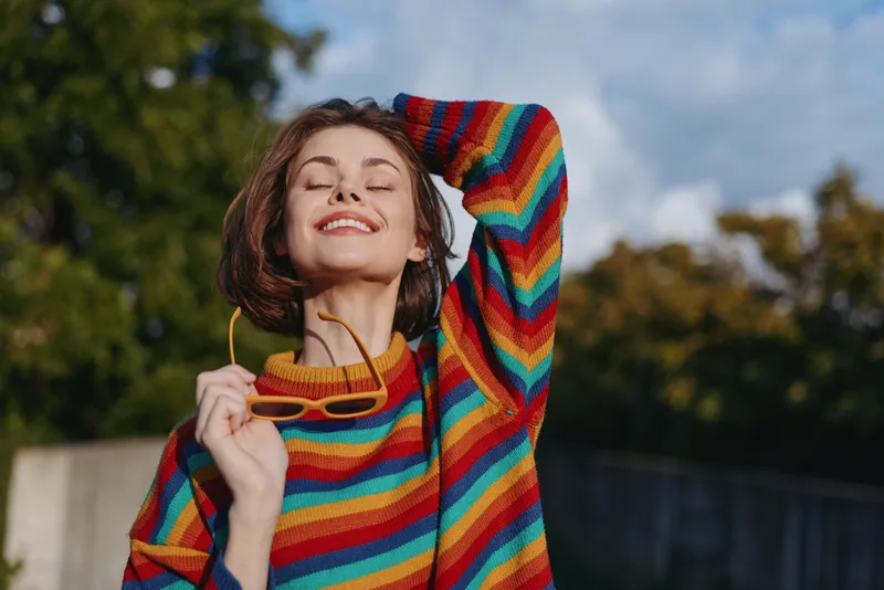 woman striped sweater sunglasses smiling young woman casual outfit short hair outdoor joyful portrait of a 20s female model with eyes closed, hand behind head, colorful knit in sunny park lifestyle