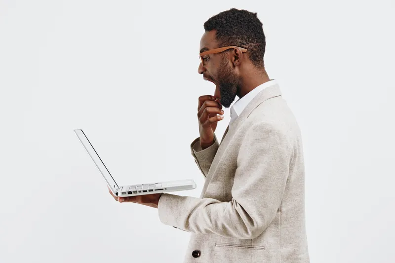 Thoughtful man holding laptop and contemplating, dressed in beige blazer and glasses, isolated on plain white background, studio portrait. Business and technology concept