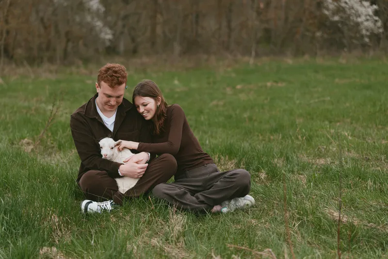 Cheerful couple outdoors, sitting on grass, gentle moment of affection as they cuddle a small white pet, soft sunlight, relaxed mood, natural tones, peaceful park scenery
