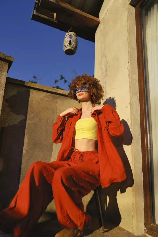 Cheerful woman outdoors in a bold red jacket and yellow top, relaxed pose on a sunny terrace, natural lighting, vibrant lifestyle moment and carefree vibe today