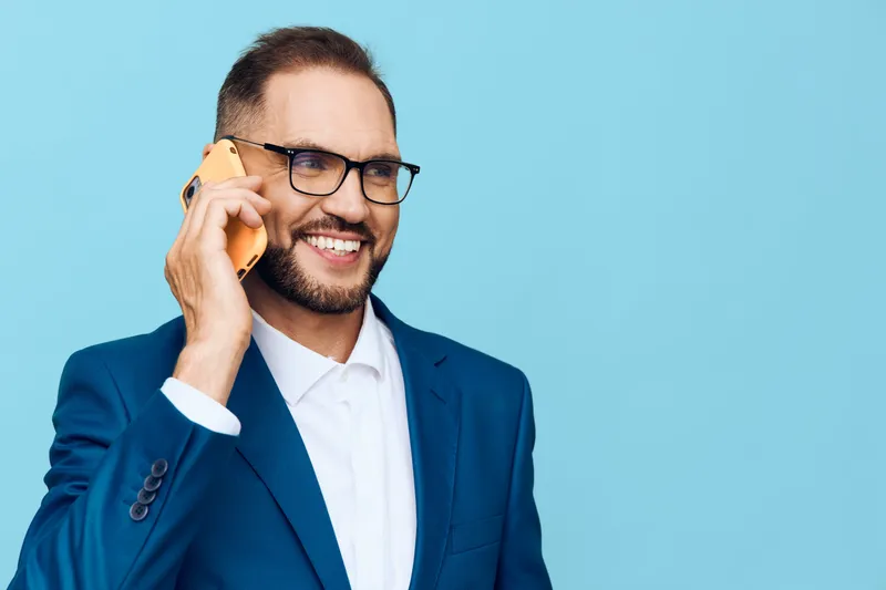 A professional man in a tailored blue suit stands against a solid blue background, confidently speaking on a mobile phone and showing warm, approachable focus and poise.