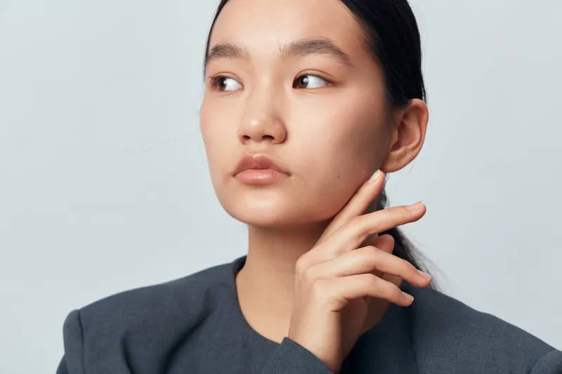 Portrait of a young woman with clear skin looking away thoughtfully wearing dark jacket on light background for beauty and fashion concept with natural makeup and hairstyle.
