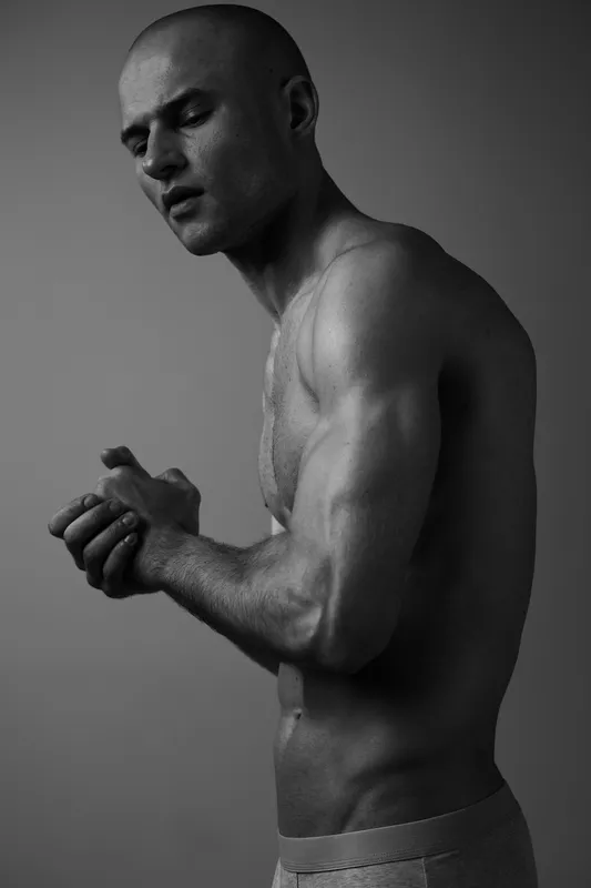 Athletic man in a thoughtful pose, showcasing his strong physique and intense expression. The image is captured in black and white, highlighting his features effectively.