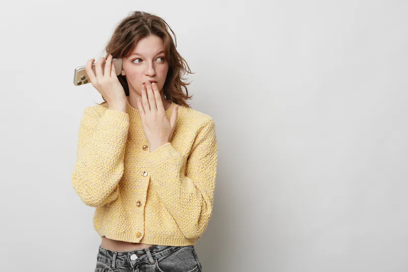 Young woman in yellow sweater holds smartphone near ear and looks surprised with hand covering mouth. Isolated on white background. Concept of unexpected news, communication, and emotion.