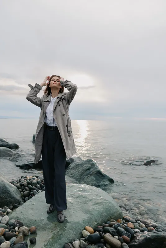 Young woman stands on rocky shore wearing coat and wide pants, looking up with hands on head. Overcast sky and calm sea create peaceful natural outdoor scene.