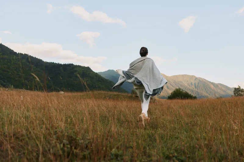 Young woman wearing a light shawl glides across a sunlit meadow with distant mountains, enjoying a carefree moment and open space
