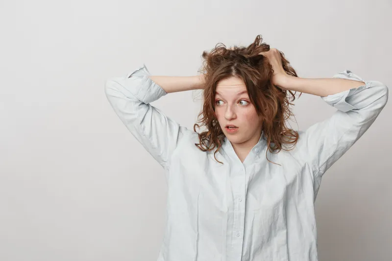 Surprised young woman with curly hair in white shirt holds head with hands, looking to side. Concept of shock, confusion, and unexpected news in casual studio setting.