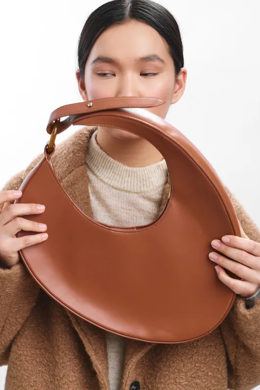 Asian woman holds a stylish bag close to her face, showcasing a soft sweater and coat. The pose conveys elegance and calm, highlighting fashion, texture, and contemporary lifestyle in a bright studio