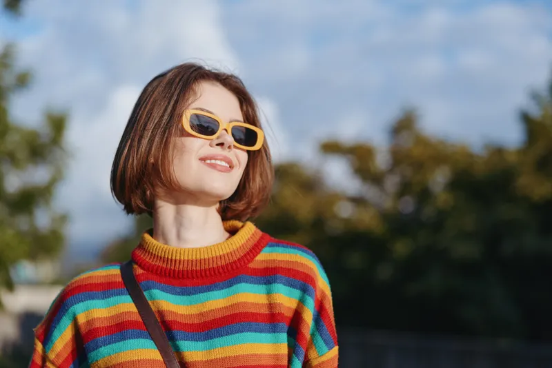woman in striped sweater and yellow sunglasses smiles while looking up outdoors, bob haircut and casual outfit in sunny city park, joyful travel lifestyle portrait with shoulder bag strap
