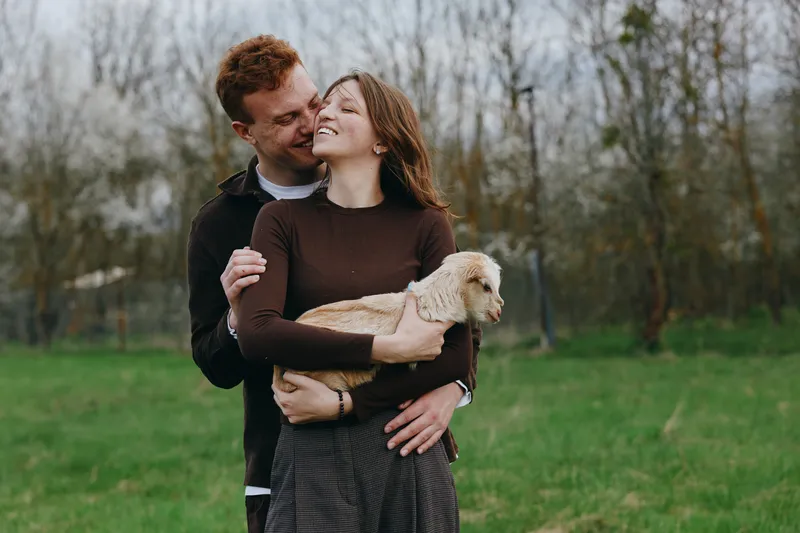 Joyful couple outdoors on a green field embrace a light brown dog, wearing cozy sweaters. Natural light highlights affection, companionship, and everyday happiness in a park setting.