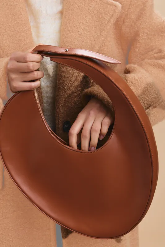 Asian woman is seen holding a circular brown handbag against a soft tan coat, showcasing a minimalist fashion accessory in a warm studio setting.