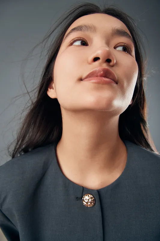 Young woman portrait with natural makeup wearing dark formal jacket against gray background. Closeup of confident female model looking slightly to side with long hair and smooth skin.