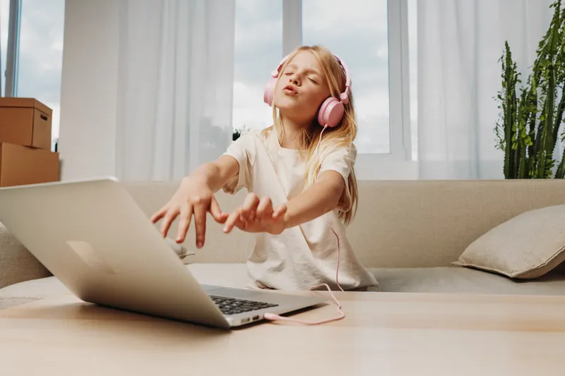 Blonde girl with pink headphones enjoys music near laptop on table in bright living room with sofa and plants. Concept of leisure, technology, and relaxation for children.