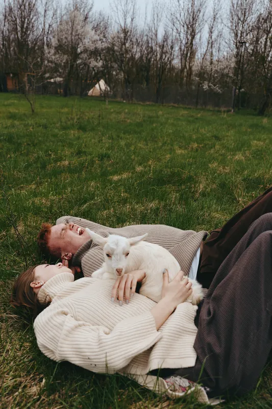 Couple relaxing on grass outdoors, affectionate embrace, white cat resting on chest, cozy sweaters, tranquil mood, natural light, casual lifestyle moment, peaceful backyard scene